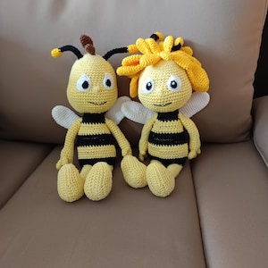 Könnte beinhalten: Zwei handgefertigte Häkelbienenpuppen. Die Bienen sind gelb mit schwarzen Streifen, weißen Flügeln und schwarzen Antennen. Eine Biene hat eine braune Antenne, die andere eine gelb-schwarze Antenne. Die Puppen sitzen auf einer braunen Oberfläche.