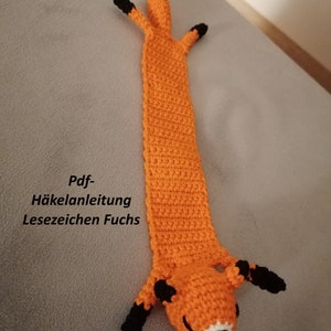 May include: Orange crocheted fox bookmark with black ears and eyes. The bookmark has the text "Pdf- Häkelanleitung Lesezeichen Fuchs" on it.