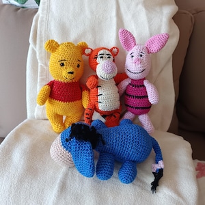 Könnte beinhalten: Handgefertigte gehäkelte Winnie-Puuh-Figuren: Winnie, Tigger, Ferkel und Eeyore. Winnie ist gelb mit einem roten Hemd, Tigger ist orange, Ferkel ist rosa und Eeyore ist blau. Die Spielzeuge werden auf einer weißen Decke präsentiert.