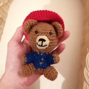 Könnte beinhalten: Ein brauner gehäkelter Teddybär, der einen roten gehäkelten Hut und einen blauen gehäkelten Pullover trägt.