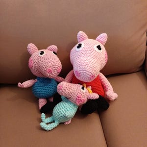Pode incluir: Três bonecas de porco de crochê feitas à mão em rosa, azul e verde-azulado. As bonecas têm olhos pretos e usam roupas de cores diferentes. As bonecas estão sentadas em uma superfície marrom.