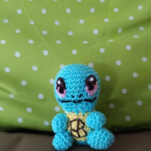 Könnte beinhalten: Handgefertigtes, gehäkeltes Squirtle-Plüschtier. Das Spielzeug ist hauptsächlich hellblau mit einem gelben Bauch und einem schwarz-gelben Schilddesign. Es hat schwarze Augen mit rosa Akzenten und einen schwarzen Mund. Der Hintergrund ist ein grüner Stoff mit weißen Tupfen.
