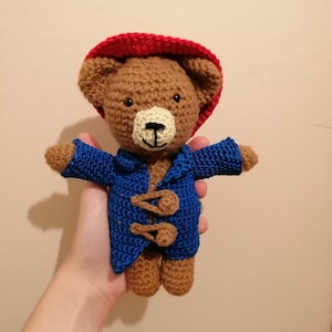 Könnte beinhalten: Ein brauner Häkelteddybär, der einen roten Hut und einen blauen Mantel mit braunen Knöpfen trägt.