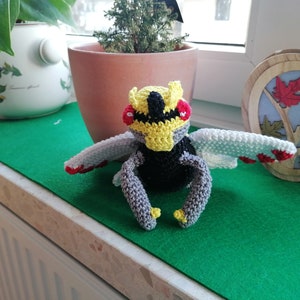 Pode incluir: Um brinquedo de pelúcia de crochê de um personagem Pokemon, com detalhes amarelos, pretos, brancos e vermelhos. O brinquedo está sentado em uma superfície verde.