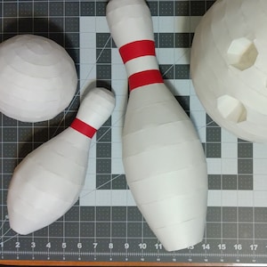 Puede incluir: Tres bolos de papel maché blanco y una bola de bolos. Dos de los bolos tienen rayas rojas. La bola de bolos tiene tres agujeros.