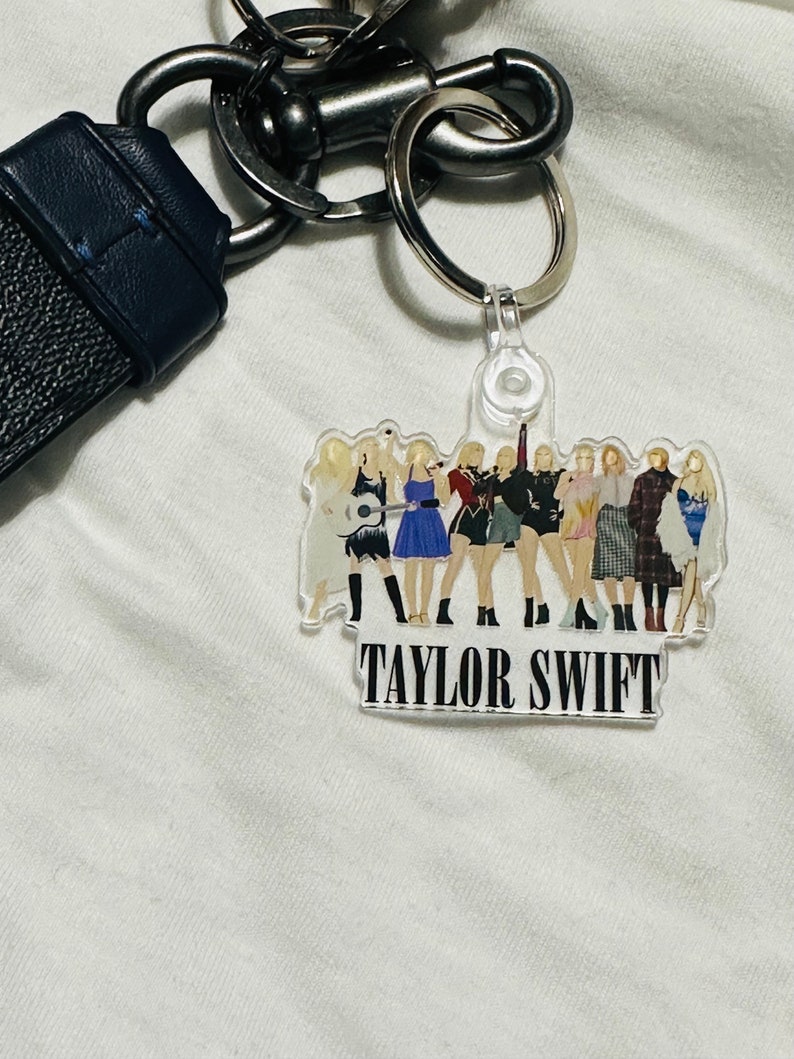 K&ouml;nnte beinhalten: Ein durchsichtiger Schl&uuml;sselanh&auml;nger aus Kunststoff mit einer Gruppe von Frauen in verschiedenen Outfits, darunter der Text "TAYLOR SWIFT". Der Schl&uuml;sselanh&auml;nger ist an einem silbernen Schl&uuml;sselring befestigt.
