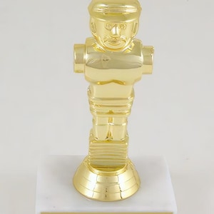 May include: A gold-colored trophy featuring a stylized figure of a hockey player. The trophy has a white marble base with a gold-colored plate.