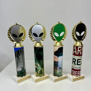 May include: Four gold alien head trophy awards with different colored alien heads and different images on the base. The alien heads are silver, green, black, and white. The images on the bases are a mountain, a green alien, a UFO, and a red and white sign that says "Warning Restricted Area Trespassing".