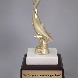 May include: A gold-colored shark trophy on a white marble base with a dark wood base. The base has a gold plaque with the text "You're gonna need a bigger boat."