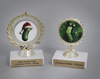 Puede incluir: Dos trofeos dorados con bases blancas. Uno presenta un pepinillo con un gorro de Papá Noel, el otro un adorno de pepinillo. Los trofeos tienen el texto "Smith Holiday Party Christmas Pickle - Winner" y "Christmas Pickle - Winner".