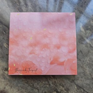 May include: A square, textured paper with a watercolor design in shades of pink and orange. Small gold flecks are scattered across the surface. The text "-Designed by Tempestt" is printed in a cursive font at the bottom.