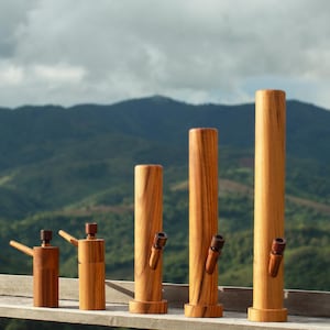 Puede incluir: Una colección de bongs de madera de varios tamaños, dispuestos sobre una superficie de madera. Los bongs son cilíndricos con un acabado de vetas de madera natural. Cada bong presenta una boquilla y un cuenco. El fondo muestra una vista panorámica de montañas verdes bajo un cielo nublado.