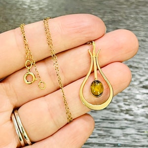 Puede incluir: Un collar de cadena de oro con un colgante con un dise&ntilde;o en forma de l&aacute;grima con una piedra de ojo de tigre marr&oacute;n.
