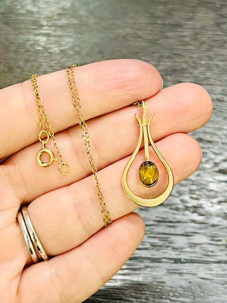 Puede incluir: Un collar de cadena de oro con un colgante con un dise&ntilde;o en forma de l&aacute;grima con una piedra de ojo de tigre marr&oacute;n.