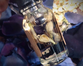 May include: A clear glass bottle with a black cap filled with a light yellow liquid and white flower buds. The bottle is sitting on a bed of purple and blue flower petals.