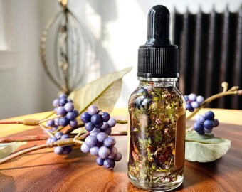 May include: A clear glass bottle with a black dropper top filled with a herbal infused oil. The bottle is sitting on a wooden surface with purple berries and green leaves.