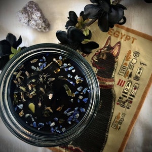 May include: A black glass candle jar with a black wax candle inside. The candle is filled with small blue, green, and gold pieces. The jar is sitting on a fabric surface with the word "EGYPT" printed on it.