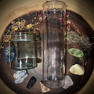 May include: Two clear glass jars, one tall and one short, on a wooden surface with dried herbs and crystals. The jars are empty and ready to be filled.