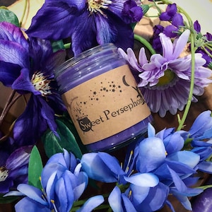 May include: Purple candle in a glass jar with a brown label that says "Sweet Persephone". The label has a black cauldron and stars. The candle is surrounded by purple flowers.