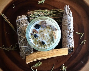 May include: A white candle in a glass jar with dried herbs, black crystals, and wood pieces. The candle is surrounded by bundles of sage, palo santo wood, and rosemary.