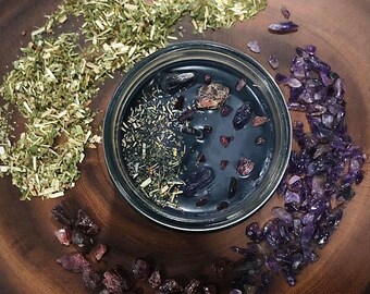May include: A black jar filled with a dark liquid and herbs, surrounded by purple and red crystals on a wooden surface.