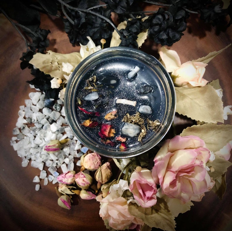 May include: A black candle with herbs, crystals, and rose petals embedded in the wax. The candle is in a clear glass jar and is surrounded by white and pink flowers.