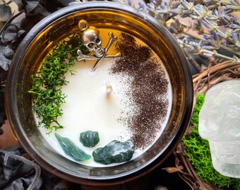 May include: A candle in a brown glass jar, with a white wax interior and a silver skull and crossbones charm. The candle is decorated with green moss, dark brown glitter, and green gemstones, creating a unique aesthetic.