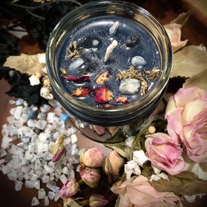 May include: A dark blue candle in a glass jar with a white wick. The candle is decorated with dried flowers, herbs, and small stones. The jar is surrounded by white stones and dried rose buds.