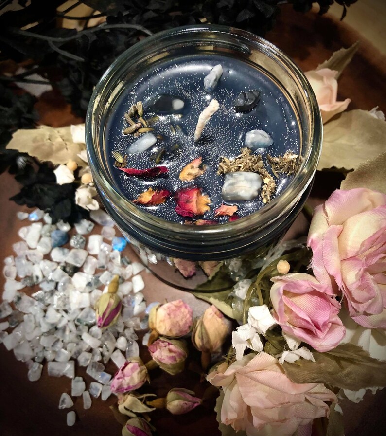 May include: A dark blue candle in a glass jar with a white wick. The candle is decorated with dried flowers, herbs, and small stones. The jar is surrounded by white stones and dried rose buds.