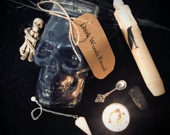 May include: A black skull-shaped glass jar candle with a brown label that reads "Dark Woods Ritual". The candle is surrounded by various witchy items, including a white candle with a black crow design, a silver spoon, a white crystal pendulum, and a small black rock.