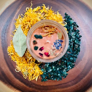 May include: A small glass jar candle with a peach-colored wax base. The candle is decorated with dried flowers, herbs, and green and blue crystals. The candle is surrounded by dried yellow petals and green crystals.