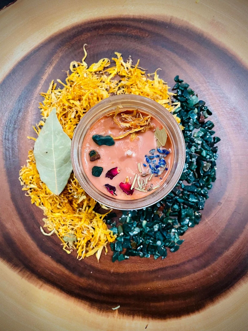May include: A small glass jar candle with a peach-colored wax base. The candle is decorated with dried flowers, herbs, and green and blue crystals. The candle is surrounded by dried yellow petals and green crystals.