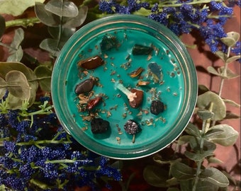 Könnte beinhalten: Tealfarbene Kerze mit eingebetteten Kristallen und Kräutern in einem Glasgefäß. Die Kerze ist von grünem und blauem Laub umgeben.