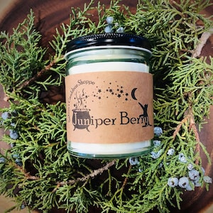 May include: A glass jar candle with a brown label that says "Salem's Cauldron Shoppe" and "Juniper Berry". The label has a black and white illustration of a cauldron with stars and a crescent moon.