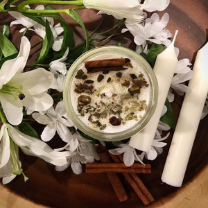 May include: A white candle with a clear glass jar filled with herbs, spices, and cinnamon sticks. The candle is surrounded by white flowers and cinnamon sticks.