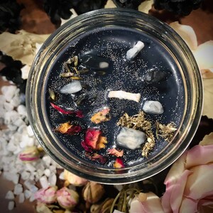 May include: A black candle with embedded crystals, dried herbs, and rose petals in a clear glass jar.