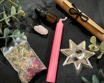May include: A pink candle, a small bag of dried herbs, a pink crystal, a small bottle of oil, a rolled up piece of paper tied with twine, and a silver star-shaped candle holder.
