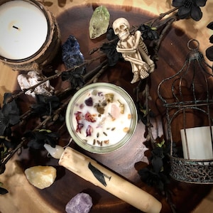 May include: A white candle with various crystals and dried flowers inside a glass jar. The candle is surrounded by other crystals, a skeleton figurine, and a birdcage with a white candle inside. The background is a wooden surface.