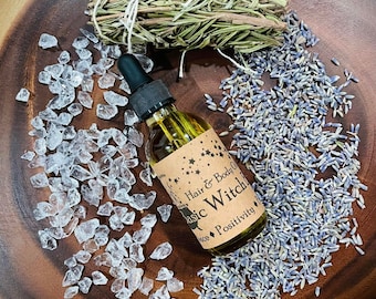 Peut inclure: Une bouteille d'huile en verre avec un compte-gouttes, étiquetée "Hair & Body Oil - Basic Witch!", est posée sur une surface en bois, entourée de cristaux clairs, de lavande séchée et d'un bouquet d'herbes noué avec de la ficelle.