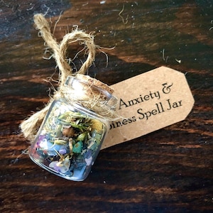 May include: A small glass jar filled with herbs, crystals, and other natural elements. A brown paper tag is attached to the jar with the text "Anxiety & Stress Spell Jar".