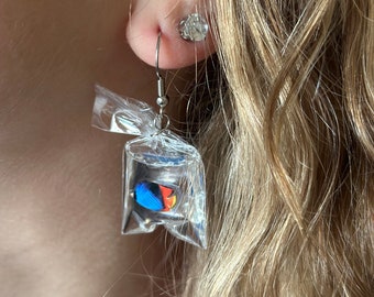 May include: A dangle earring featuring a miniature clear plastic bag filled with liquid and a small blue, black, and orange fish figurine. The charm hangs from a silver-toned metal hook wire, creating a realistic aquarium effect in a tiny package.