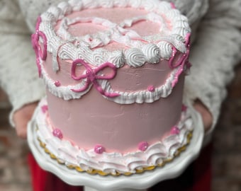 May include: A pink frosted cake with white frosting and pink bow decorations. The cake sits on a white pedestal cake stand with a gold trim. The cake is round and has a decorative border.