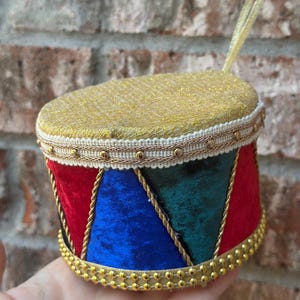 May include: A cylindrical drum ornament featuring a gold glitter top, a white and gold beaded trim, and alternating panels of red, blue, and dark green velvet. The base is decorated with a gold beaded border and includes a gold hanging loop.