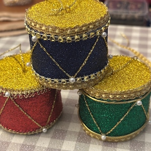 May include: Three cylindrical drum ornaments featuring glittery surfaces in red, dark blue, and green. Each drum has a gold glitter top, gold trim, and small white bead accents along the edges with gold cord lacing in a triangular pattern.