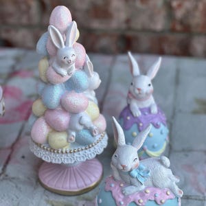 May include: Easter-themed figurines featuring white rabbits. One figurine is a pastel-colored tree with rabbits, and the others are rabbits atop decorated eggs. The figurines have a glittery finish and are in shades of pink, blue, and yellow.