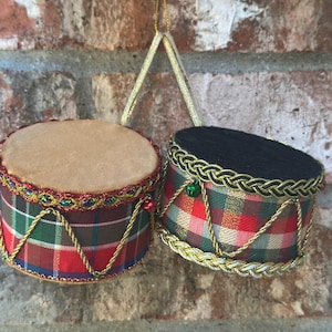 May include: Two miniature drum ornaments featuring red, green, and blue plaid fabric sides. One drum has a tan top and the other a black top. Both are decorated with gold braided trim, gold cord lacing, and small green and red jingle bells.