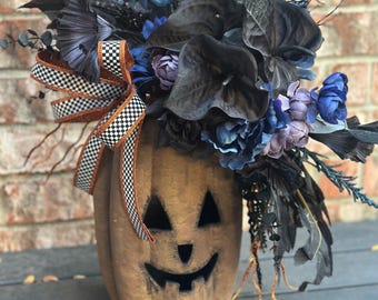 Puede incluir: Una calabaza de color canela con una cara tallada, adornada con un arreglo de flores artificiales azul oscuro y negras, follaje y una cinta decorativa con un patrón de cuadros blanco y negro con bordes de purpurina naranja.