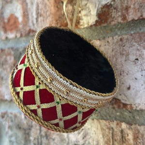 May include: A cylindrical drum-shaped ornament featuring a deep red velvet body with a gold lattice pattern and small green and red accents. The top is covered in black fabric, and the edges are trimmed with gold braided cord and white ribbon.