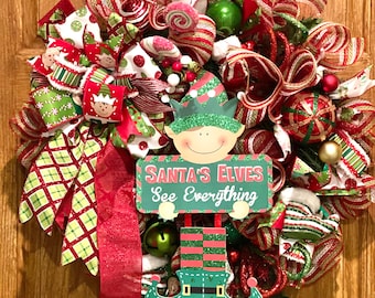 May include: A festive Christmas wreath with red, green, and white ribbons, ornaments, and an elf figure holding a sign that reads "Santa's Elves See Everything." The wreath is displayed on a wooden door.