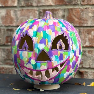 May include: A decorative pumpkin with a carved jack-o'-lantern face, featuring a hand-painted pattern of overlapping circular brushstrokes in vibrant shades of purple, teal, pink, light blue, white, and metallic gold across the entire surface.